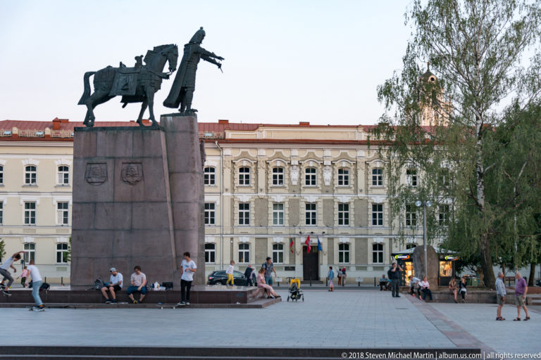 King Mindaugas equestrian monument on a tall pedestal in a public square with seated visitors and historic buildings behind.