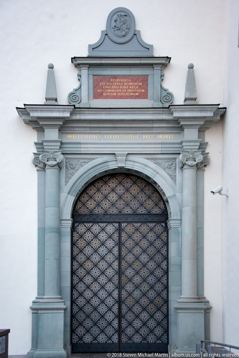 Closed patterned metal doors within an ornate arched stone gateway topped by inscriptions and heraldic decoration.