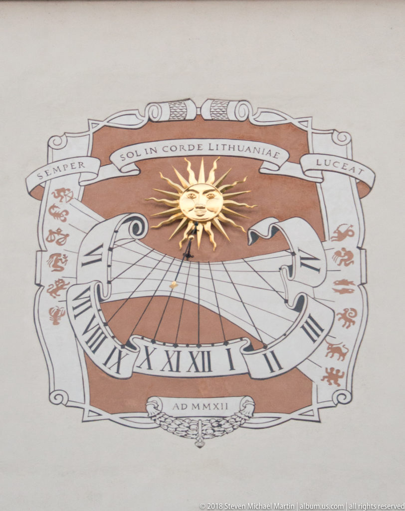 Decorative wall sundial with Roman numerals, projecting gnomon, golden sun, heraldic motifs, and Latin inscription.