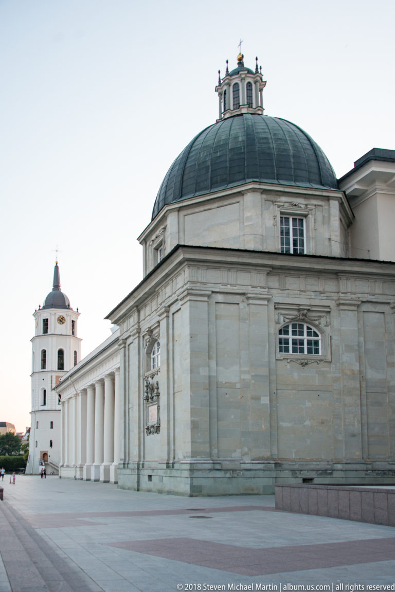 Side view of Vilnius Cathedral with colonnade, large domed chapel, and the freestanding bell tower visible beyond.