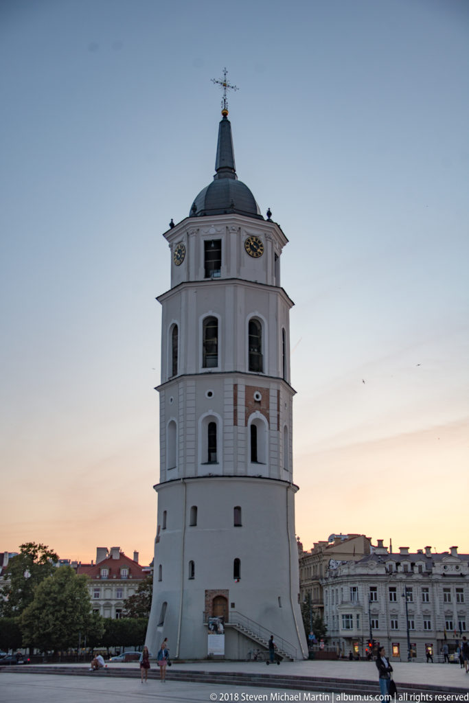 Freestanding white Vilnius Cathedral Bell Tower with clock faces, tiered stages, dark roof, and pedestrians at its base.