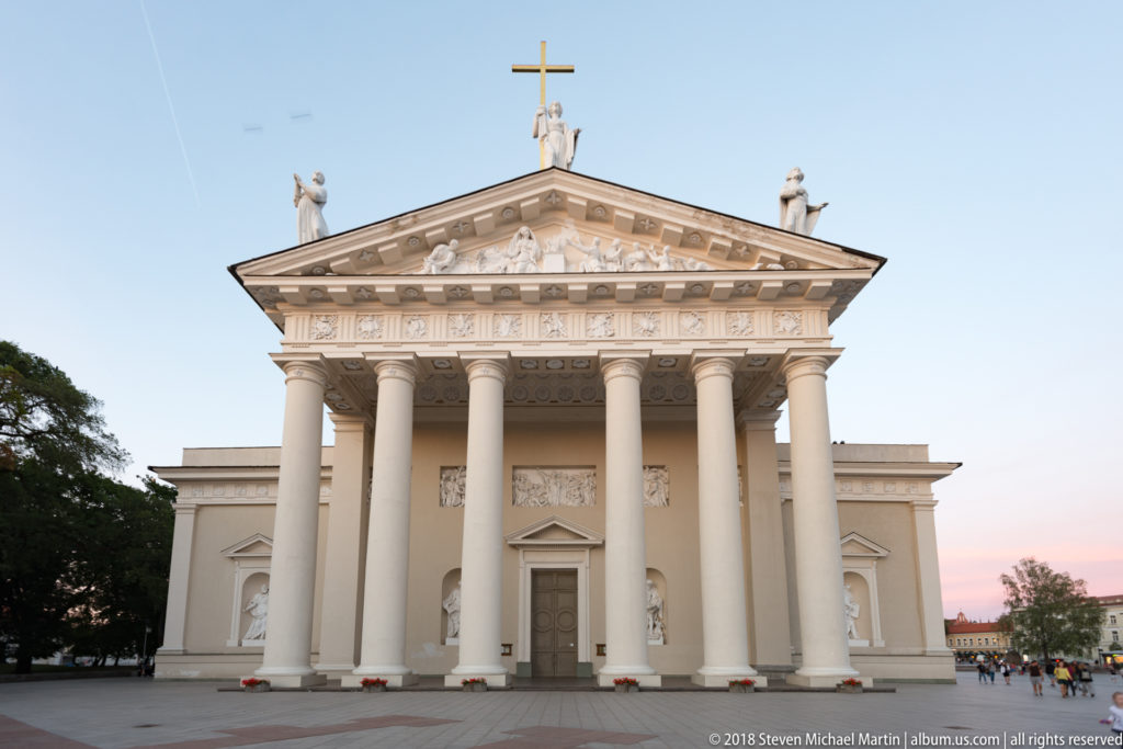Frontal view of Vilnius Cathedral with six-column portico, sculpted pediment, rooftop statues, and broad open square.