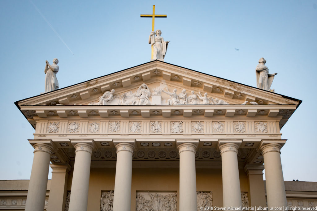 Close upward view of Vilnius Cathedral’s classical columns, sculpted triangular pediment, rooftop statues, and cross.
