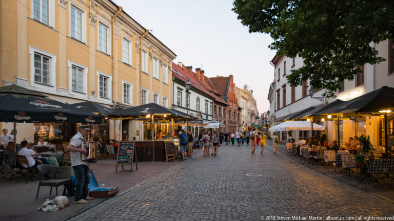 Cobbled Vilnius Old Town street lined with outdoor cafés, awnings, historic façades, and evening pedestrians.