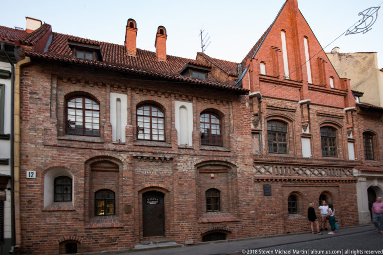 Red-brick Gothic building with pointed gable, arched windows, decorative masonry, and white plaster accents in Vilnius Old Town.