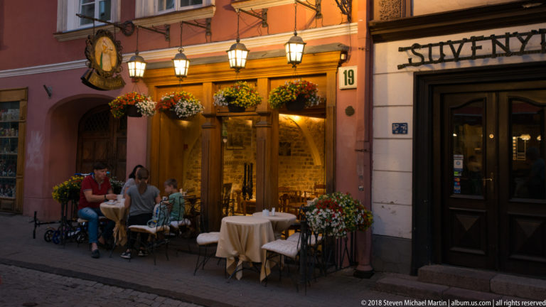 Warmly lit restaurant entrance with hanging flowers, outdoor tables, diners, lanterns, and building number 19.