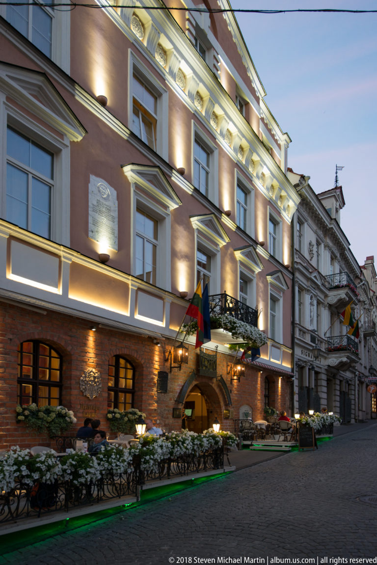 Illuminated historic building with flags, flower boxes, arched openings, and outdoor seating along a cobbled street.