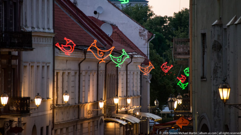 Narrow Vilnius Old Town street at dusk with colorful bird-shaped lights suspended above illuminated historic façades.
