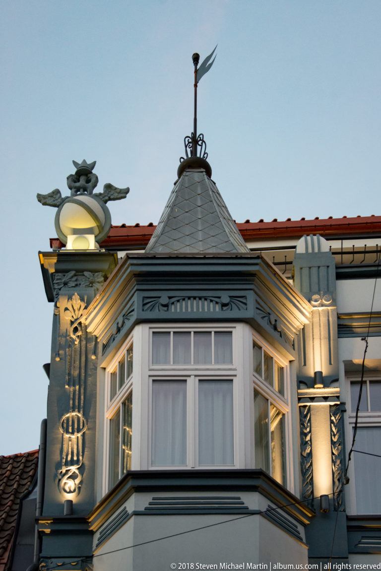 Illuminated projecting corner bay with tall windows, carved trim, steep roof, and whimsical rooftop ornament.