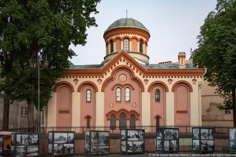 Front of a pink Orthodox church with arched doorway, paired windows, decorative brickwork, and a tall domed drum.