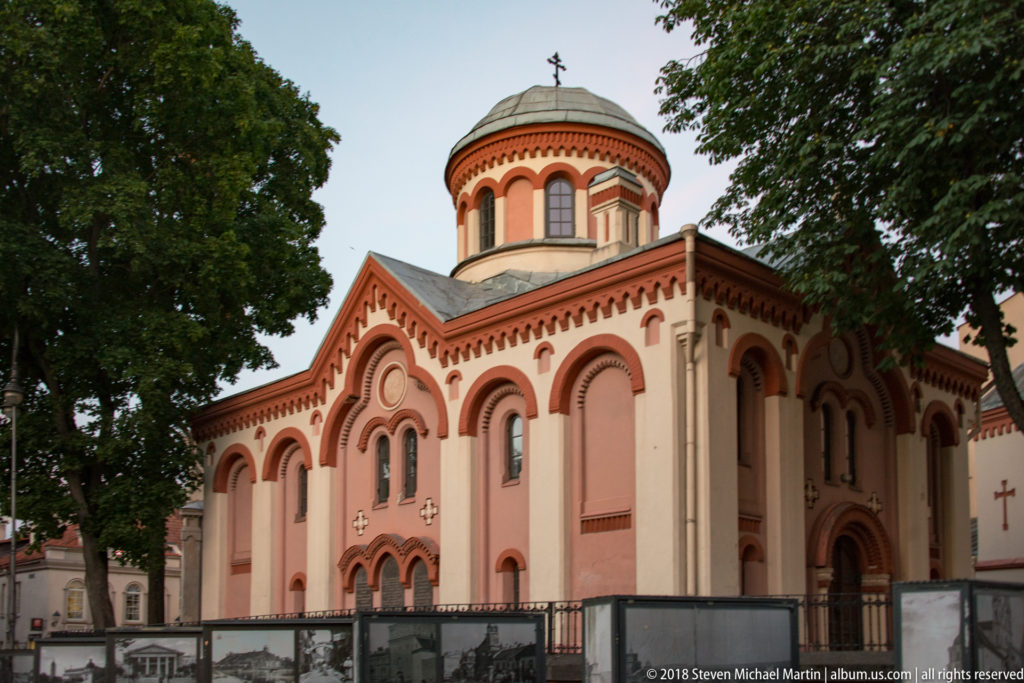 Pink Orthodox church with rounded arches, decorative brick bands, and a broad domed drum framed by trees.