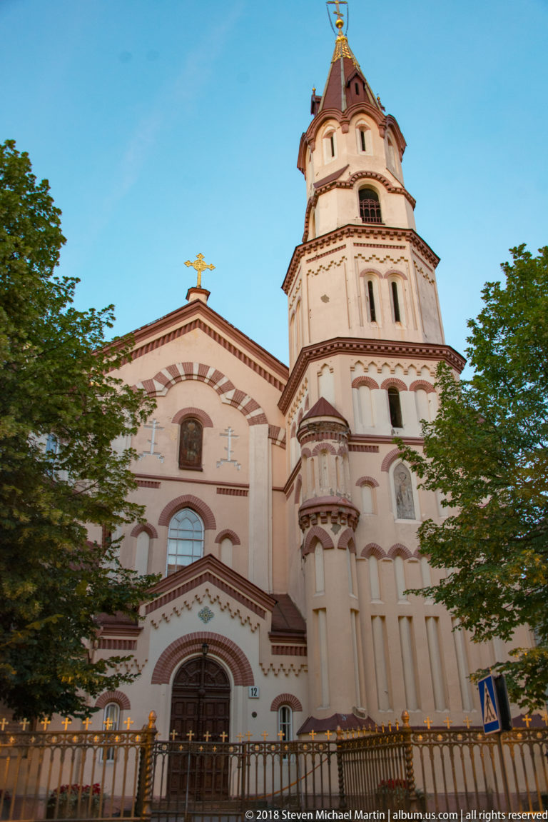 Decorative pale church façade with adjoining tiered bell tower, arched openings, and contrasting trim above trees in Vilnius.