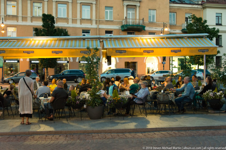 Crowded outdoor café terrace with striped awnings, diners, potted plants, and historic façades in central Vilnius.