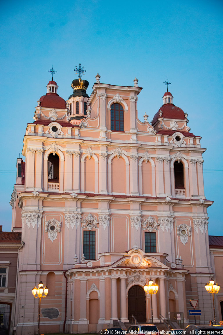 Centered dusk view of St. Casimir’s pink Baroque façade with pilasters, domed side towers, and crown-shaped feature above.