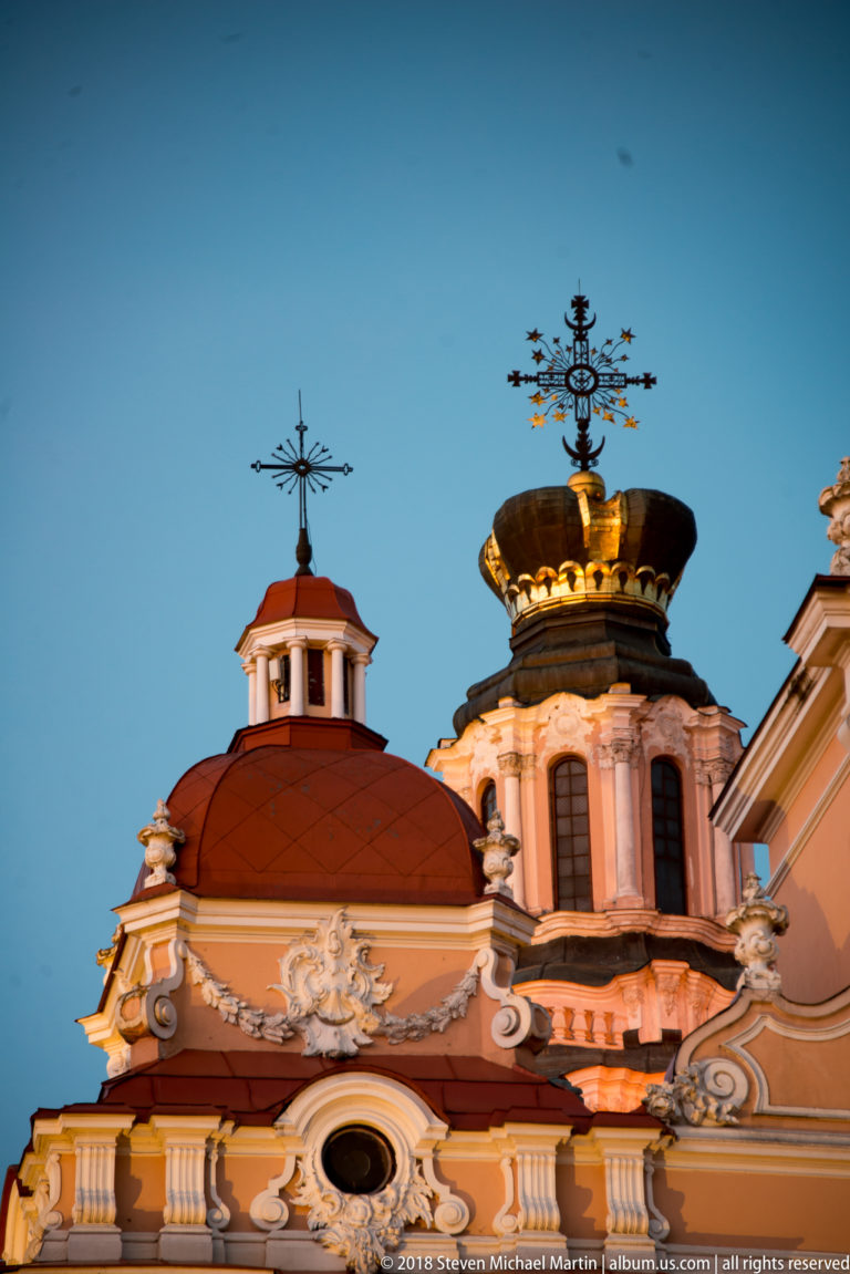 Crown-topped dome, red roof lantern, white Baroque ornament, and crosses of St. Casimir Church against blue sky.