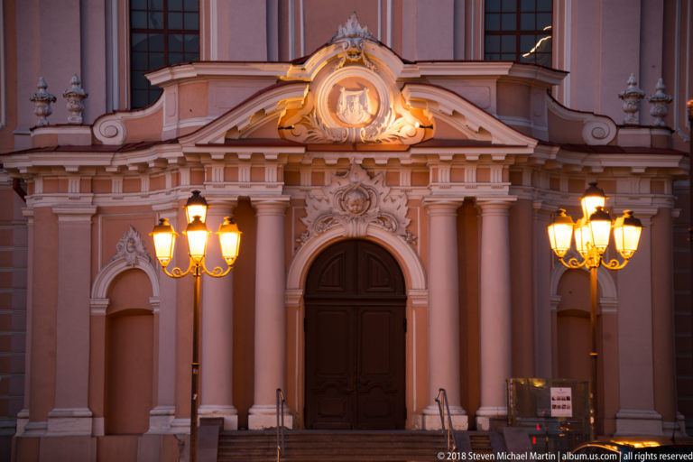 Close view of St. Casimir’s Church entrance with dark wooden doors, paired columns, carved ornament, and glowing street lamps.