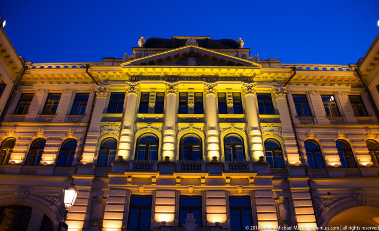 Grand illuminated historic façade with central columns, triangular pediment, arched windows, and rusticated masonry at night.