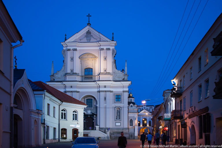 White Baroque church façade at the end of an Old Town street, with a separate arched religious gateway at left.