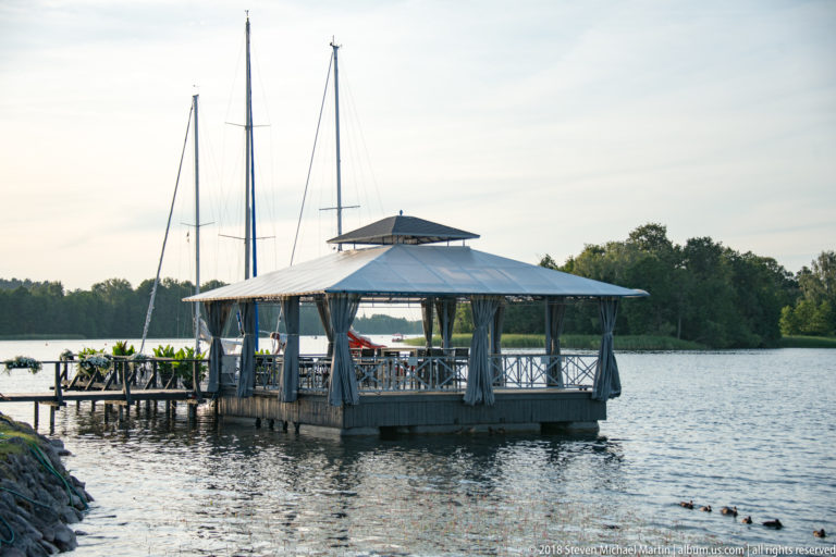 Roofed open-air pavilion over Lake Galvė with tables, chairs, dock, and sailboat masts behind.