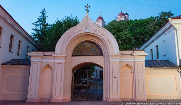 Pale pink religious gateway with a painted sacred scene above the arch, metal gates, and domed church towers behind.