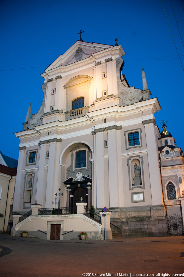 Illuminated pale Baroque façade of St. Theresa’s Church with pilasters, niches, cornices, and a central arched entrance.