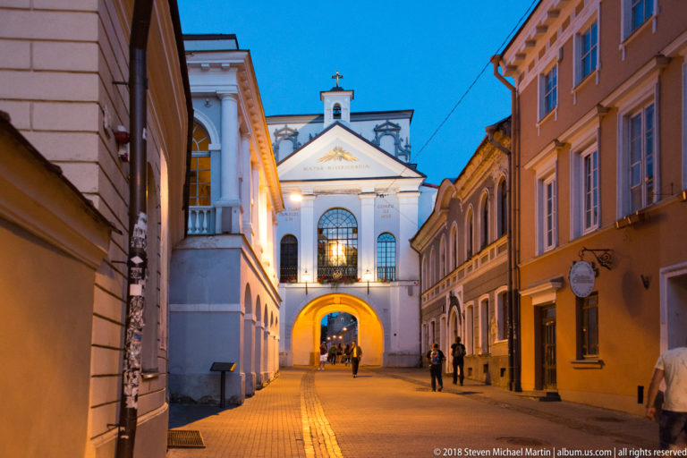 Gate of Dawn at the end of a narrow evening street, with pedestrians passing through the lit archway below the chapel.