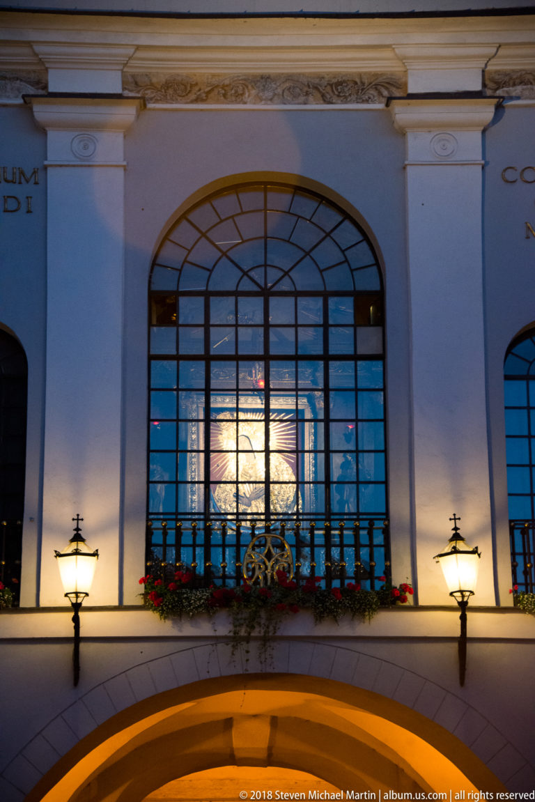 Large arched window glowing above the Gate of Dawn passage, with ironwork, flowers, and wall lamps at night.