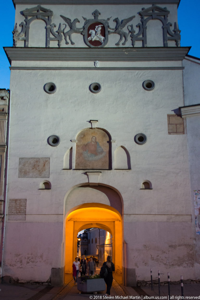 White Gate of Dawn façade with glowing arched passage, religious panel above, and heraldic crest near the roofline.