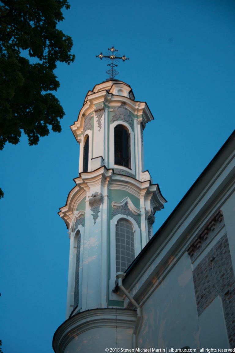 Pale Baroque bell tower with arched openings and curved cornices rising above a church roof at dusk.
