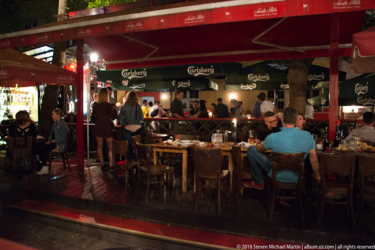 Crowded outdoor restaurant terrace at night with diners seated at wooden tables beneath large branded awnings.