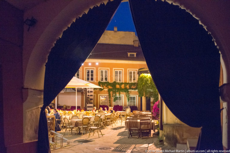 Dark curtains beneath an arch frame a brightly lit courtyard café with rows of outdoor tables and chairs.