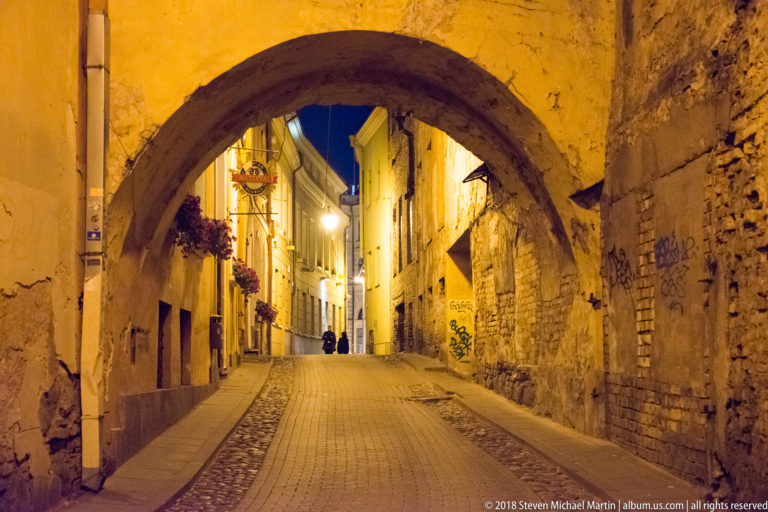 Large worn masonry arch framing a narrow uphill Old Town lane with cobbles, lamps, and hanging flowers at night.