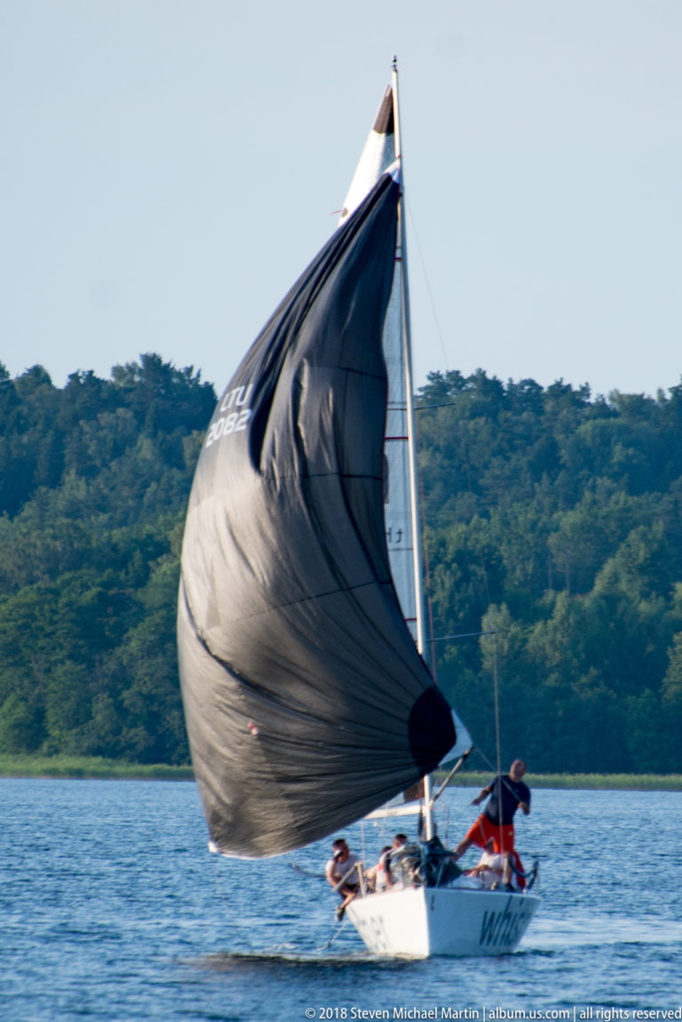 Small sailboat carrying a large dark spinnaker across Lake Galvė, with two sailors aboard and wooded shore behind.