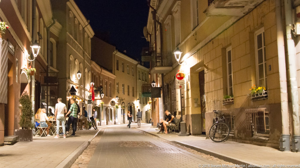 Narrow Vilnius Old Town street at night with outdoor diners, pedestrians, bicycles, and illuminated historic façades.