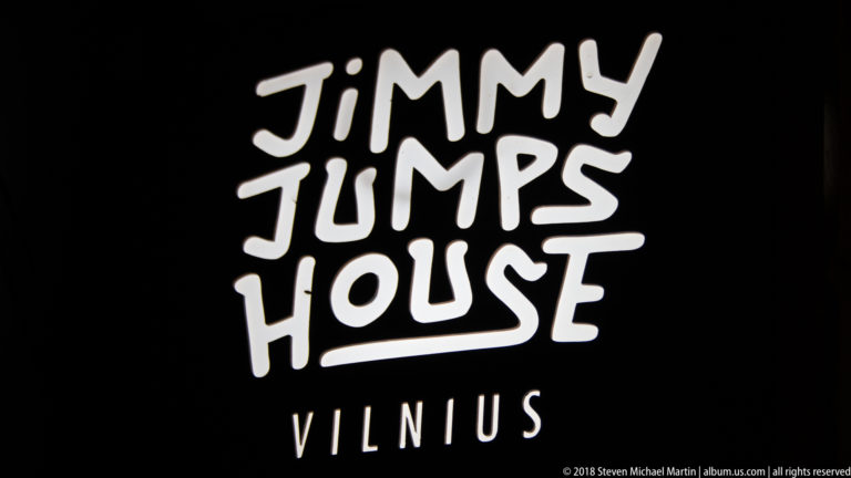 Black sign with large white hand-lettered words “JIMMY JUMPS HOUSE” and smaller “VILNIUS” beneath.