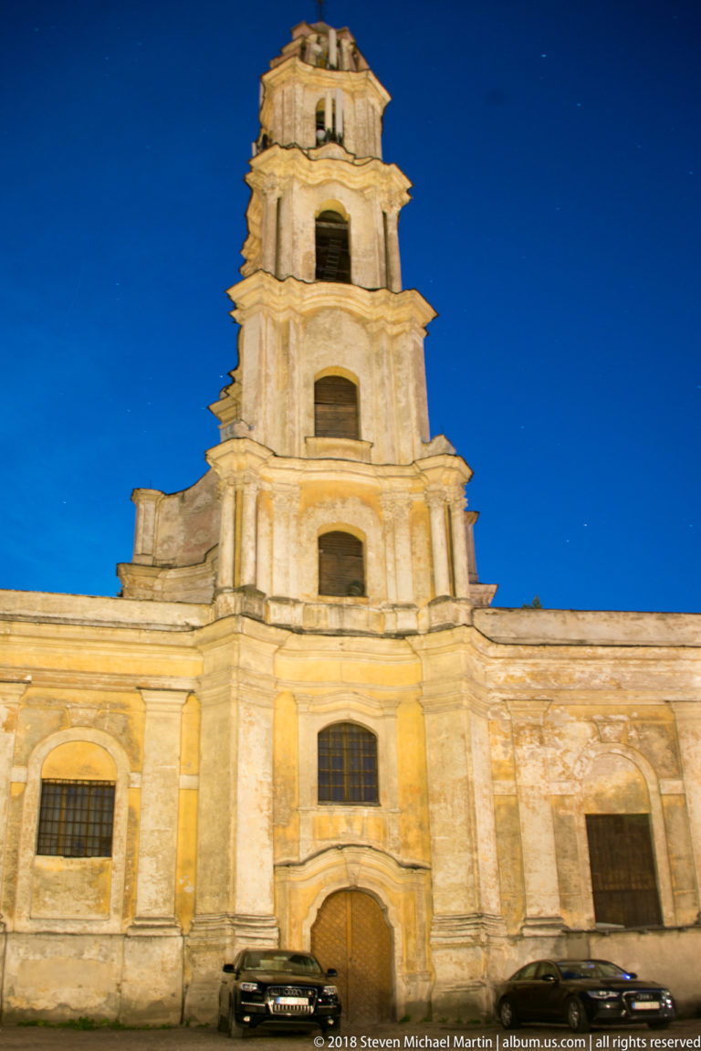 Weathered pale church bell tower with stacked stages and arched openings, illuminated against a deep blue evening sky.