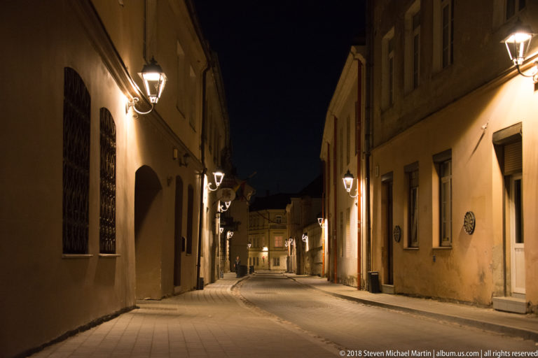 Quiet narrow lane at night with warm wall lamps, arched openings, stucco façades, and buildings receding into the distance.