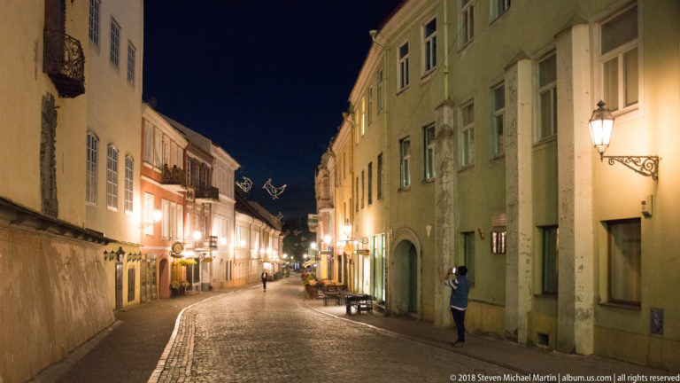 Narrow cobbled Old Town street at night, lined with pale historic buildings, lanterns, and a few pedestrians.