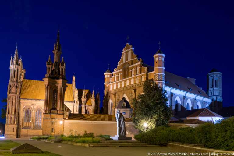 Wide night view of St. Anne’s Gothic towers and the adjoining Bernardine church across an open landscaped space.