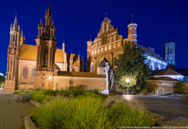 Illuminated red-brick St. Anne and Bernardine church complex behind a standing monument and landscaped foreground at night.
