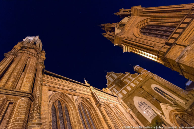 Low-angle night view between red-brick Gothic church towers with pointed windows, pinnacles, and ornate masonry.