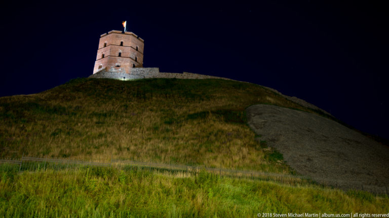 Brick Gediminas’ Tower with Lithuanian flag on the summit of a steep grassy hill at night.