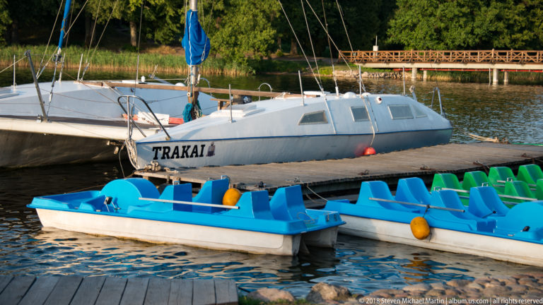 Blue and green pedal boats beside a white sailboat labeled TRAKAI at a small dock on Lake Galvė.