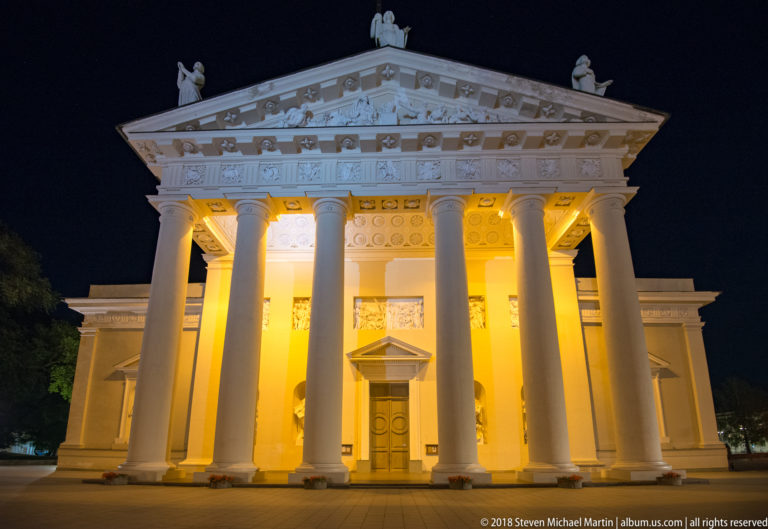 Floodlit neoclassical façade of Vilnius Cathedral with six tall columns and sculpted pediment against a dark night sky.