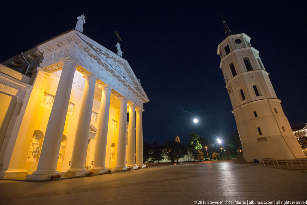 Floodlit Vilnius Cathedral portico and freestanding bell tower on Cathedral Square beneath a dark sky and bright moon.
