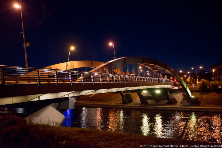 Illuminated modern bridge with curved supports and metal railings reflected in the Neris River at night.