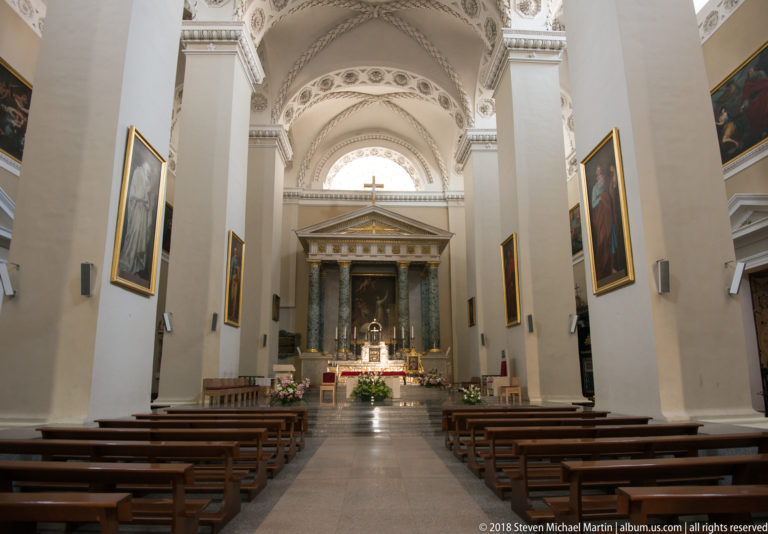 Broad cathedral nave with white piers, gilded paintings, wooden pews, and green-columned high altar ahead.
