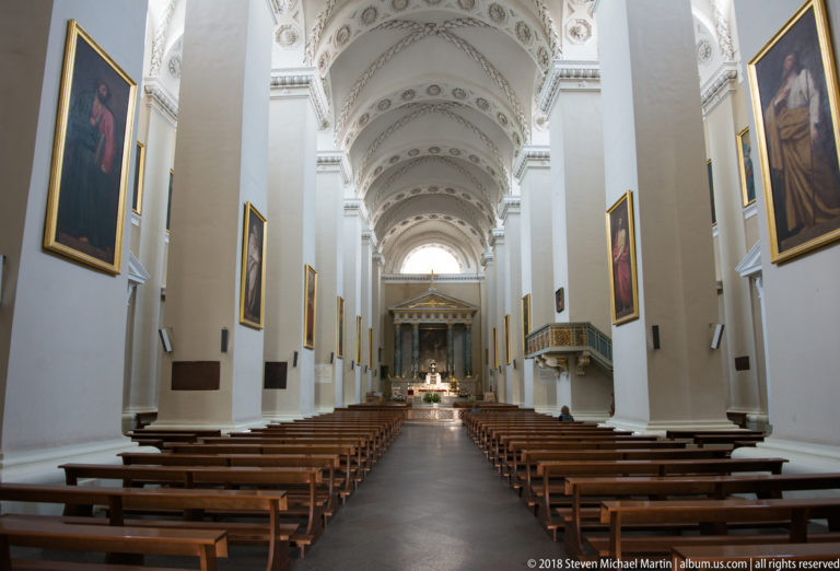 Long Vilnius Cathedral nave with wooden pews, white piers, framed paintings, vaulted ceiling, and distant high altar.