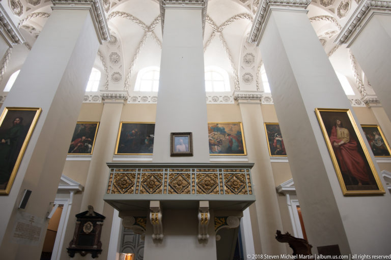 Tall white cathedral piers with gilded religious paintings and a patterned pulpit or gallery projecting between them.