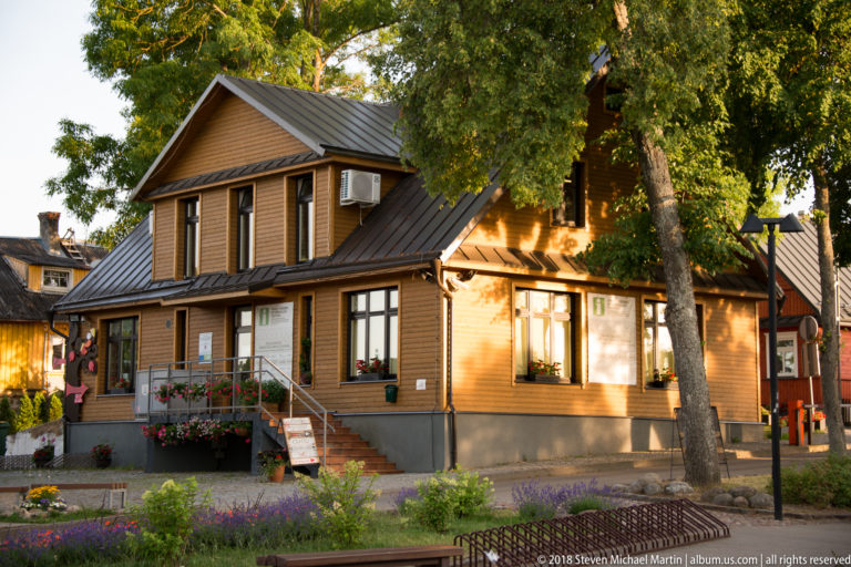 Brown wooden house with steep metal roofs, white-trimmed windows, flower boxes, and garden beneath mature trees in Trakai.
